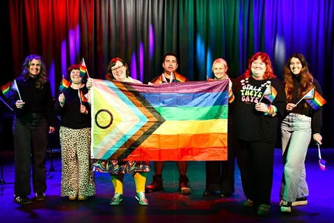 Carly Carr, Agostina Hawkins, Virginia Winter, Shannon Ross, Carol Stewart, Molly Wilder and Meg Bennett waving pride inclusion flags and smiling on the PAC stage. Photo by Nick Moseley, Portland Observer 