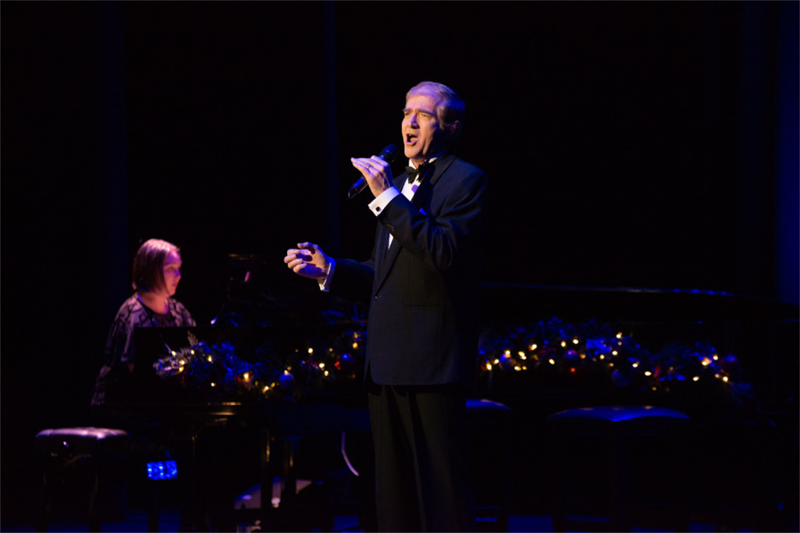 A man in a tuxedo is singing into a microhpone whlist a woman in a dark evening dress plays a grand piano decorated with Christmas lights.