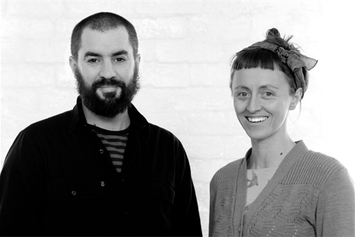 A black and white photo of a man with a beard and a woman with an edgy haircut and a scarf in a bow.