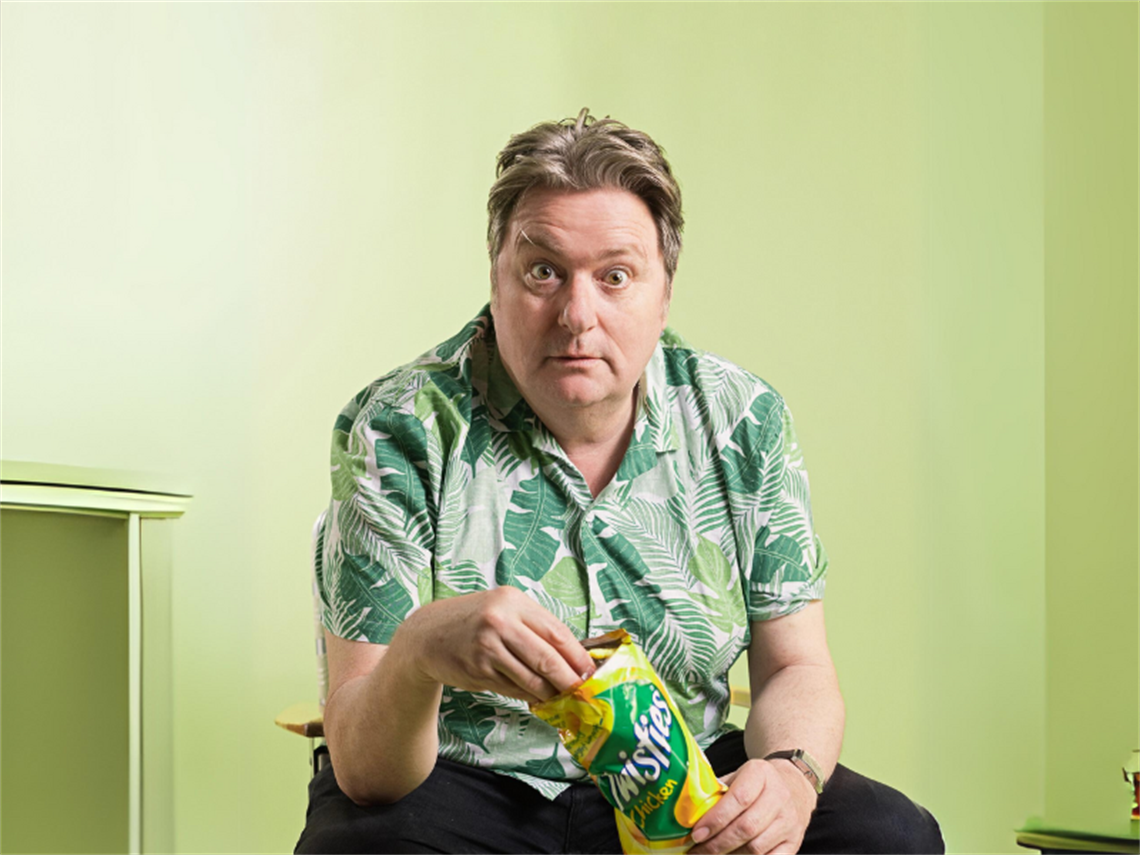 Dave Oneil wearing a green Hawaiian in a green room eating chicken flavoured twisties