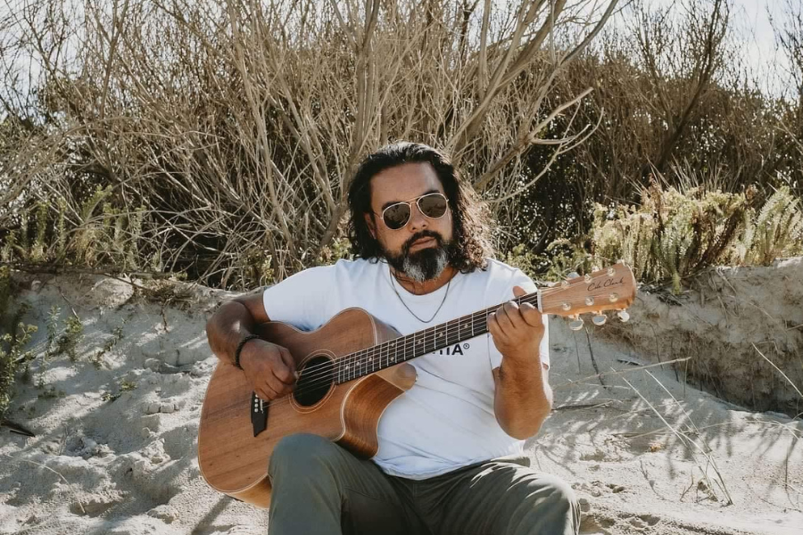 Jayden sitting on a sand dune with scrubland behind him, playing an acoustic guitar.