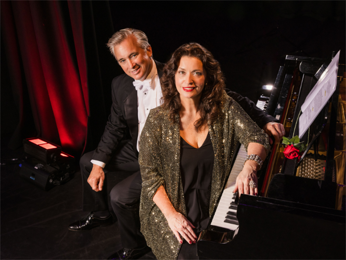 A man and a lady dressed in formal attire sitting at a grand piano with a red rose laid on top of the piano