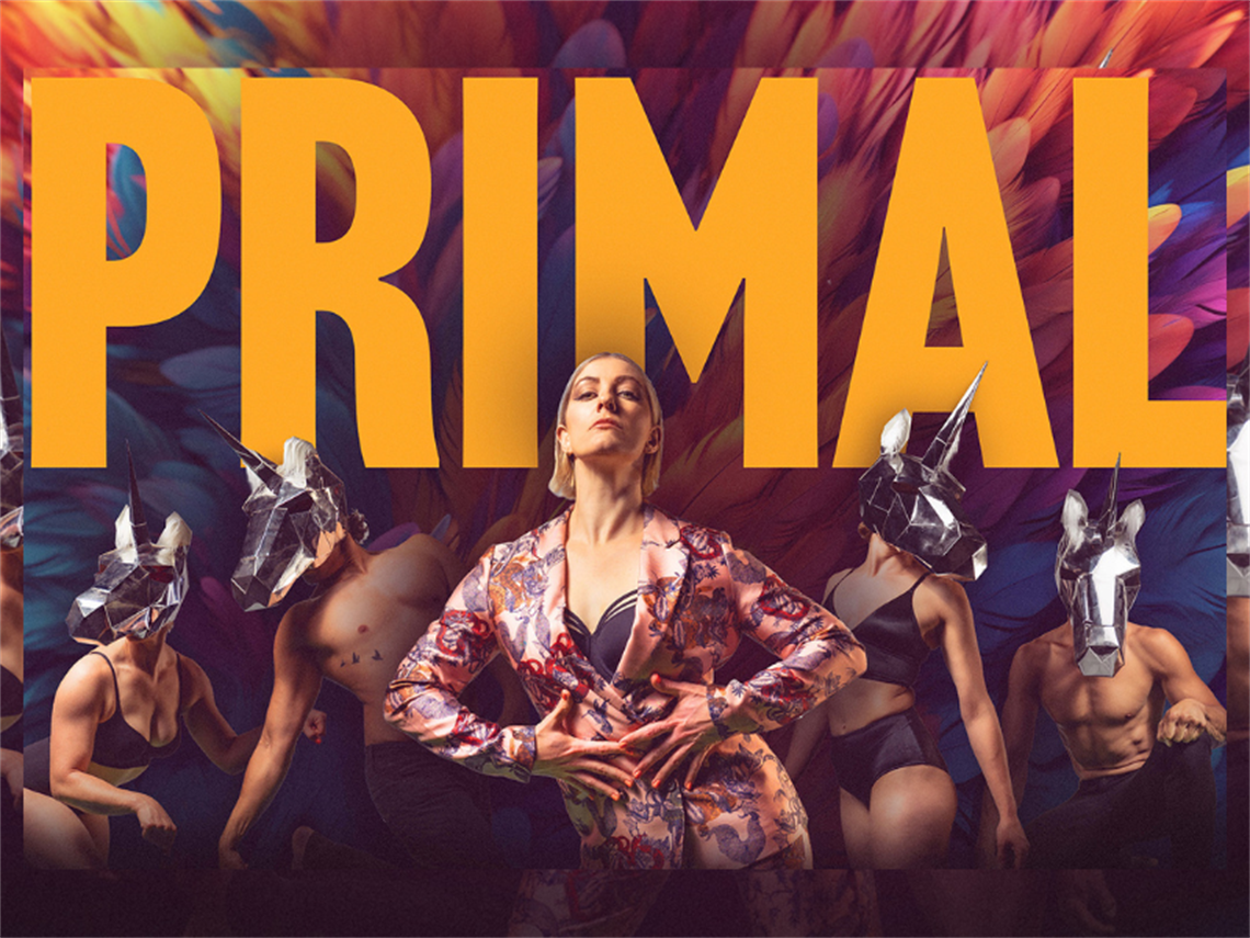several male and female acrobats in black underwear wearing silver unicorn masks behind a defiant looking young woman wearing a floral pantsuit