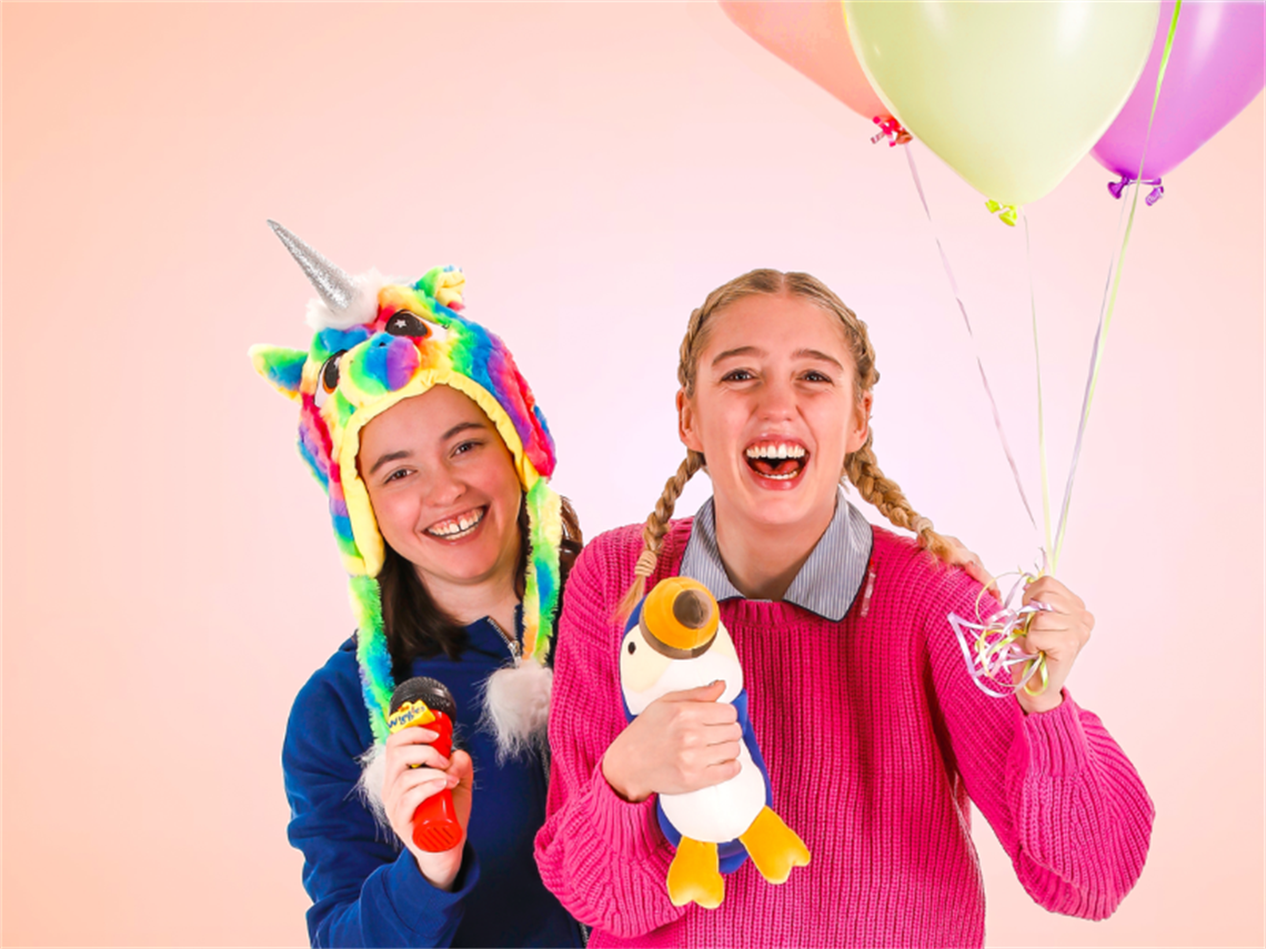 Two young girls dressed in bright colours holding soft toys and helium inflated balloons, one girl wearing a multicoloured unicorn head peice