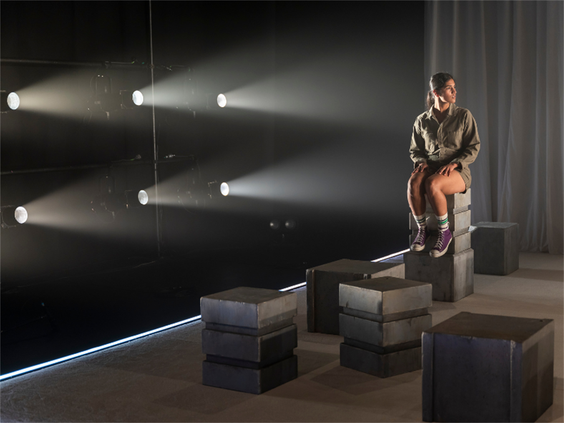 Young Girl sitting on grey blocks in a dark room with lights shining on her. Image credit for Hero Image: Brett Walker, 2022 cast ●	Image Credit for show production shots:Tiffany Garvie, 2022 cast