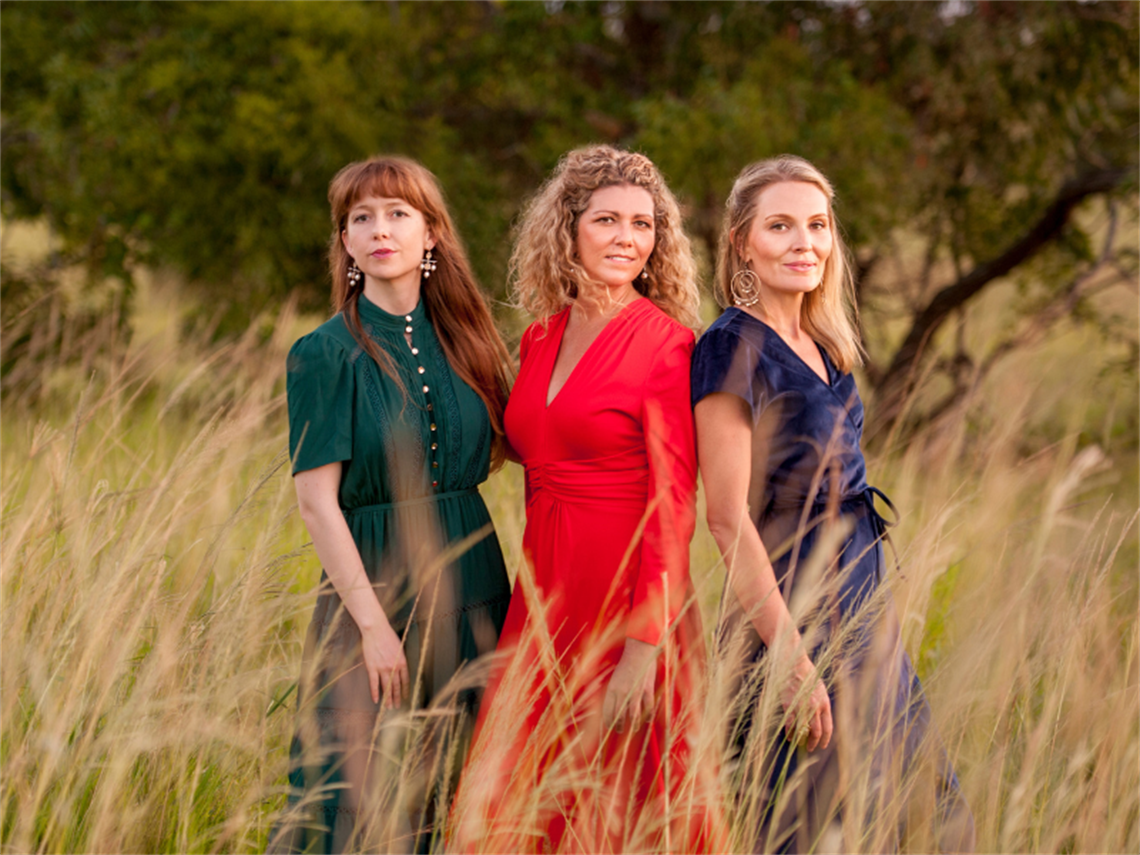 Three ladies wearing long dresses in a long dry grassy field with bush backdrop