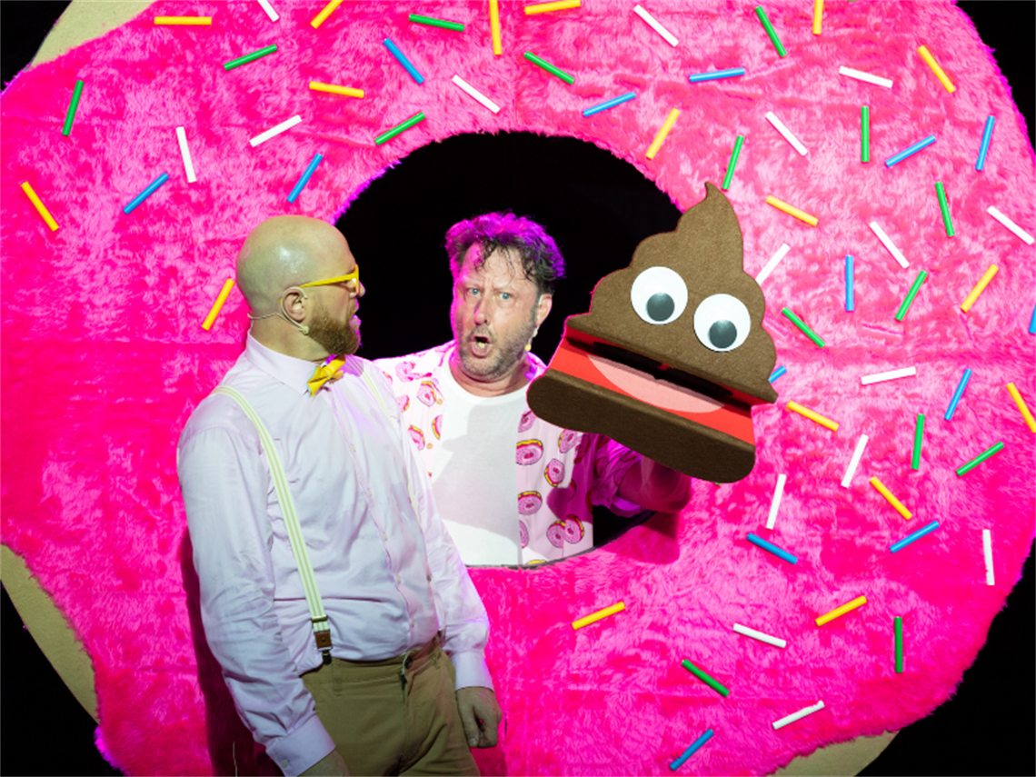 Two men dressed in pink shirts standing with a giant freckly donut and one man holding a giant emoji poo prop