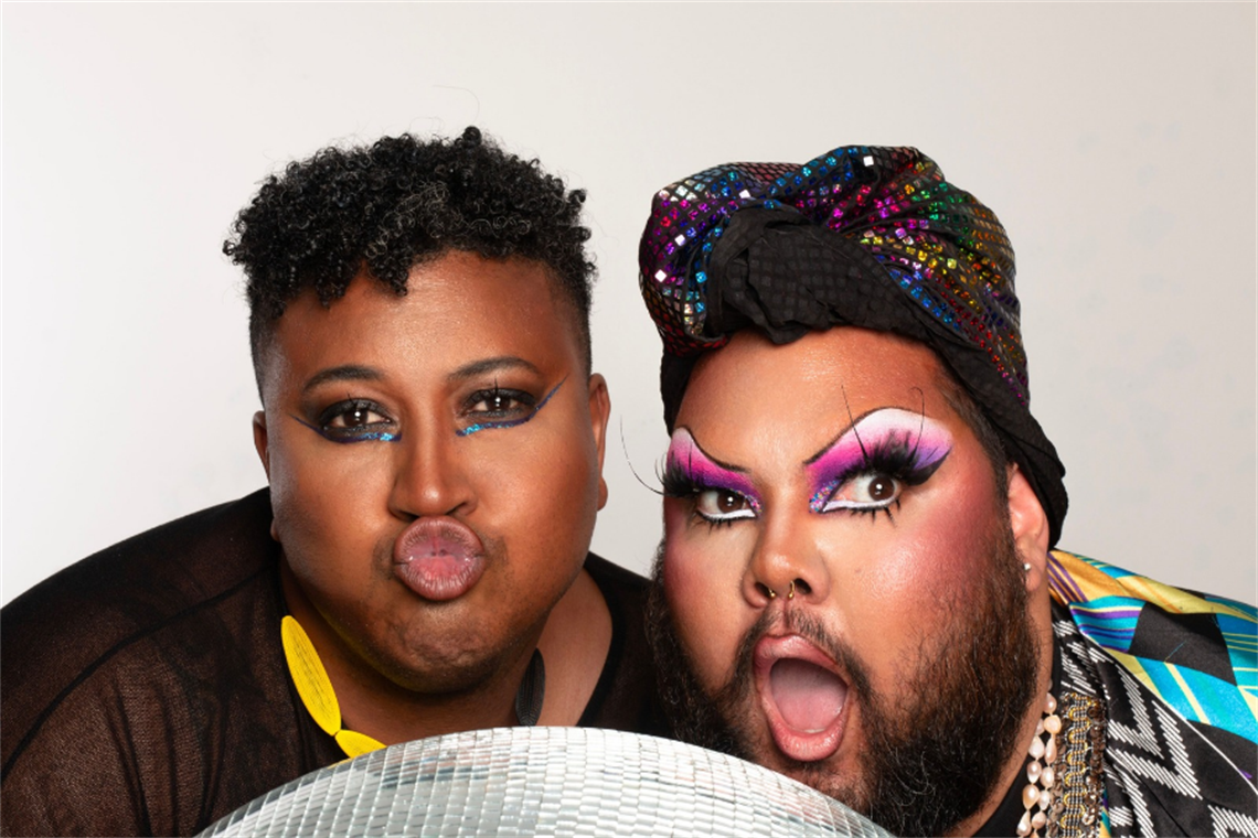 Close up of Sam Gaskin and 2Joocee in bright make up, wearing sparkly outfits, and serving up runway faces next to a mirror ball Close up of Sam Gaskin and 2Joocee in bright make up, wearing sparkly outfits, and serving up runway faces next to a mirror ball