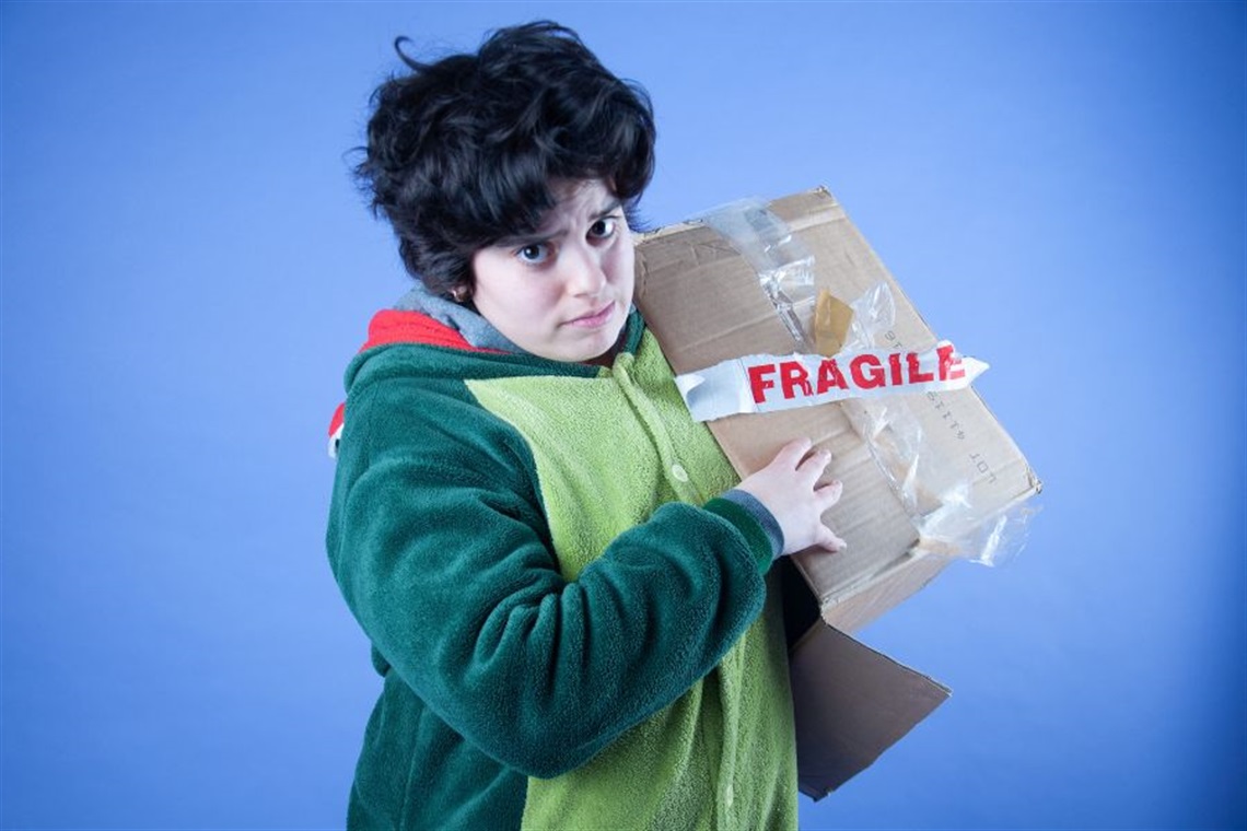 Person with short dark curly thick hair dressed in a green animal suit and holding a cardboard box with the word 'FRAGILE' written in red