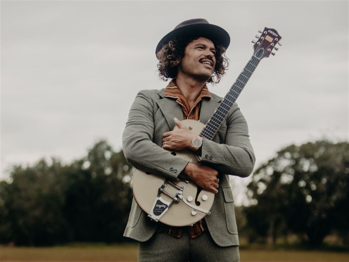 Ash Grunwald wearing a felt hat hugging a guitar in a brown and cream stripy shirt with a grey suit jacket and pants