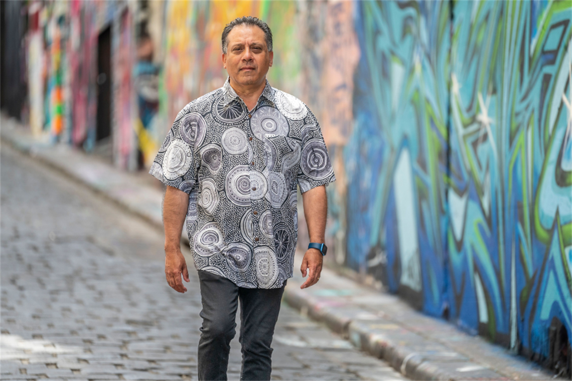 Man standing in an alley way wearing a black and white indigenous design shirt, with graffitti art on the walls of buildings behind him.