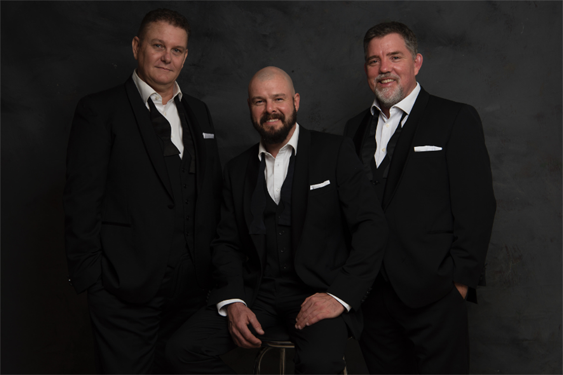 3 men smiling at the camera looking relaxed dressed in dark suits with white collared shirts unbuttoned at the top.