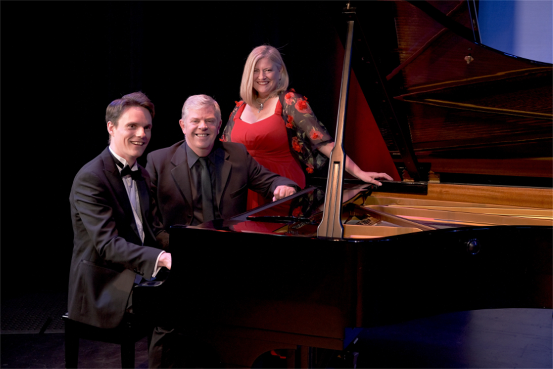 Two men sitting and one lady standing behind a piano on stage