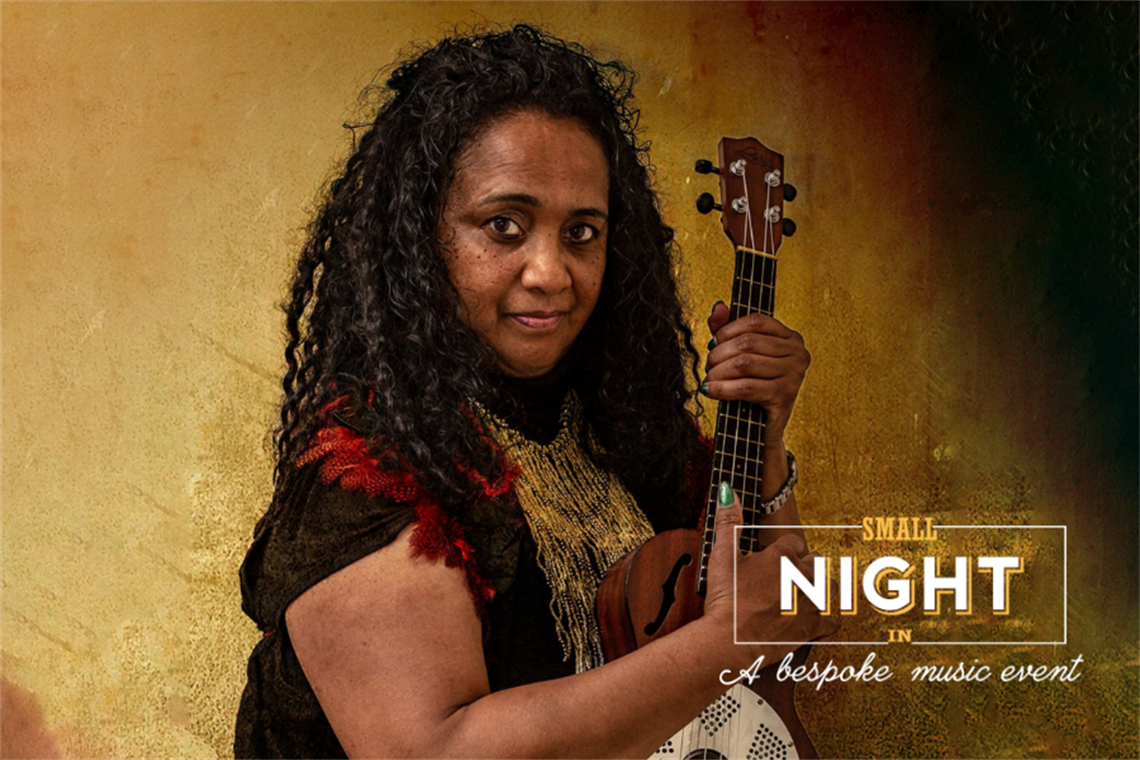 A strong looking woman with beautiful brown skin and curly dark hair stares defiantly at the camera. She is holding her resonator ukulele and wearing deep shades of black and red. Behind her is an ochre coloured wall.
