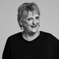 A black and white photo of Jane Clifton, a woman in her 70s with short, fair hair, wearing a black top and smiling