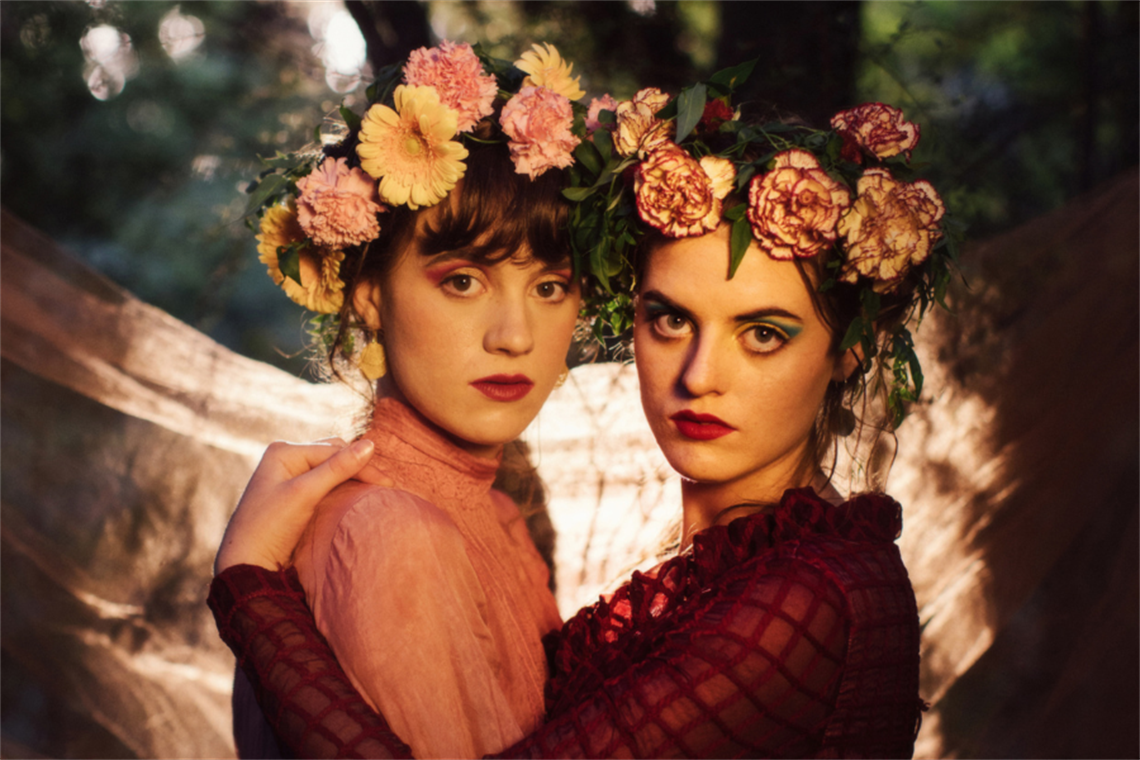Two young women with flower crowns hold each other, looking intensely into the camera