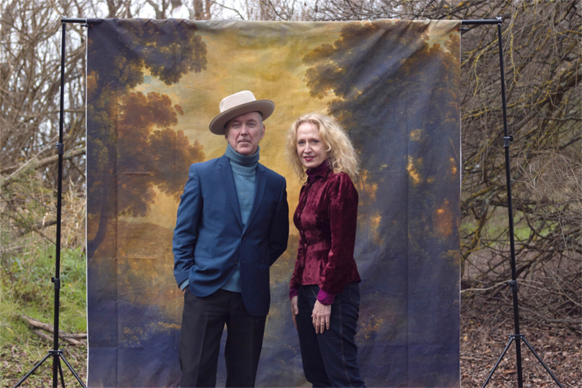 Dave Graney and Clare Moore stand in front of a painted backdrop of the Australian Bush, itself in front of real Australian bushland